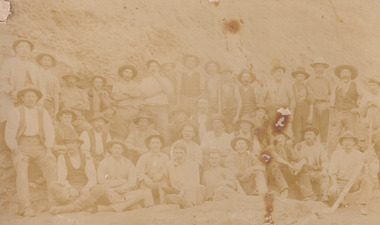 A faded sepia photograph depicting 34 men in work clothing in front of a bank of earth