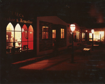 Colour photograph of  a dimly lit streetscape with a reddish hue showing a line of shops with 19th-century lamp posts. A jeweller's shop with high arched windows is in the foreground, with an image of two Victorian gentlemen visible in the window.  Source:  Burke Museum Collection.