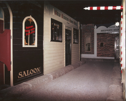 Coloured photograph containing a street of shops. Includes a saloon and hotel on the left, with a barber shop pole on the right of the image.