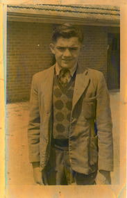 Sepia Image of a young man in a suit, in front of a building. He wears a knit vest with a geometric pattern, with a shirt and tie, trousers and a jacket.