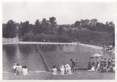Black and white photograph of people sitting on the grass by a lake watching a swimming carnival.