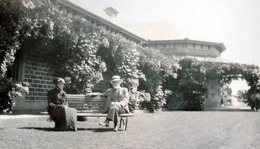 Elderly couple sitting on garden seat in front of Victorian homestead