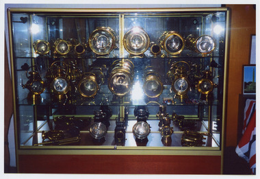 Shows a collection of coach lights that was housed in The Marysville Museum in Marysville in Victoria. The collection is displayed in a wooden cabinet with two glass shelves.
