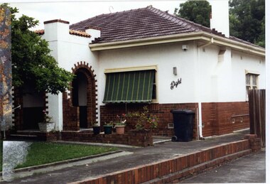 Partial front view of 2/3 rendered plus 1/3 unpainted brick house with a fortress-like central entrance porch with 2 open arches with steps, 2 tall rendered chimneys with brick features, very low brown brick side fence beside the drive on the left and low brown brick wall over the concrete path and driveway.