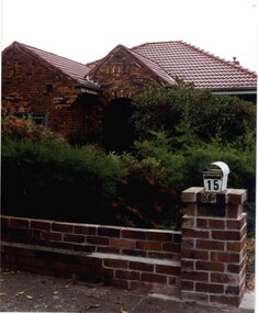 Partial front view of house made of mixed shades of brown bricks and low brick fence.  Arched porch entrance behind large bushes in the garden.  White metal letterbox on brick fence pillar.