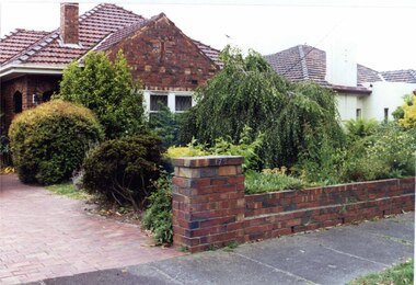 Partial front view of brown brick house with two arches into porch with a matching low brick fence.  Small trees and bushes fill the front garden with a paved drive on the left.