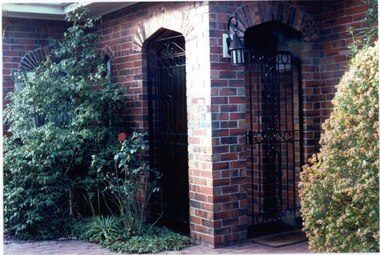 Front porch of variegated brown brick with 2 arched entries, both with closed tall black metal gates.  Door mat on brown paving outside the right entry and garden beds to the left and right filled with well-established shrubs and bushes.