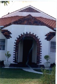 White porch with brown brick features around the open arch, floor and steps, gable and roof line of the porch reflected also on the gable of the second storey above it.  Large urns with small shrubs on either side of the concrete path leading to the porch.