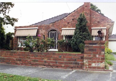 Front view of variegated brown brick house and matching low brick fence with brown metal letterbox.  It has a chimney, ornamental arch in the brickwork over the decorative white metal entry door, striped awnings over the windows, established shrubs in the garden beds and a drive on the right.