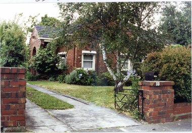 View of front and left side of variegated brown brick house with an arched porch, encased striped awnings and well-established garden beds around the house and low matching brick front fence.  Black metal letterbox behind front fence right pillar which also has a black metal gate beside the drive.