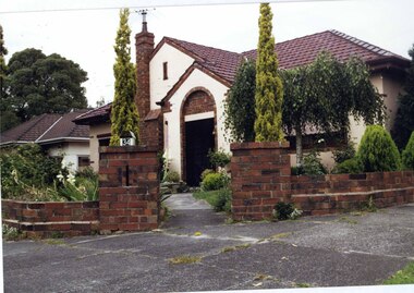 Front view of cream 2 storey home with variegated brown brick features from the street corner at the low matching brown brick fence's pillar entry.  The house has a tall brown brick chimney, wide arched porch with black metalwork door, awnings, well-established garden including tall twin pencil pines with a concrete path to the door.  Neighbouring house on the left is partially visible.