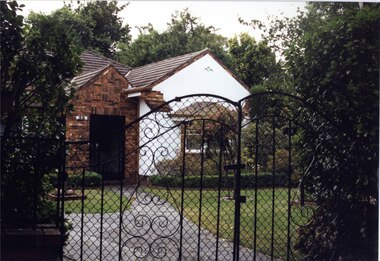 Partial front view of white house with brown brick features through decorative black metal fence.  Large brick porch with black metal door.  Well-established garden.