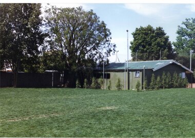 Large grassed area in front of a long green-coloured building with tall poles and fence behind that.  A couple of large trees to the left of the grass and some small trees in front of the building.