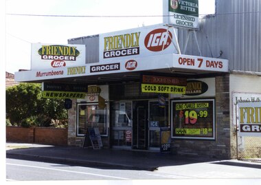 Large multi-coloured brick shop with many signs, plate glass windows, window advertising and central entry.  Four different signs say "FRIENDLY GROCER IGA".