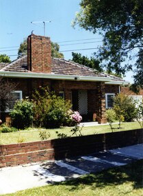 Partial front view of a mixed brown brick house with an open porch including 2 brick pillars and white trellis on the house wall, a wide tall feature brick chimney, white-framed windows and guttering matching the just visible white garage door to the right.  The established garden is behind a low brown brick fence.