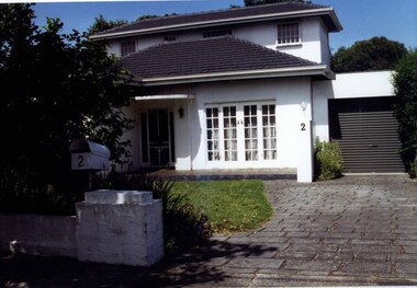 Partial view of two storey white house with adjoining roller door garage.  It has a small porch on its patio, paved drive and a brick fence painted white with a white metal letter box on its pillar.