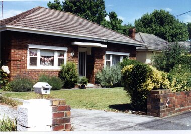 Brown brick house with small porch over the central front door, with white features on those, the windows, the guttering and the metal letterbox on the front fence.  The front garden has established plants.