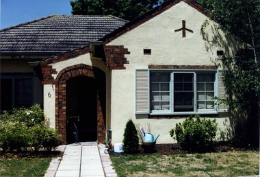 Partial view of front of cream-coloured house with brown brick features including around the arched porch entry, roof line and windows. There is a simple aeroplane-like feature above the right-hand windows.  A paved path leads through the lawn and garden beds to the front porch where there is bicycle stored.  Tall trees to the right side and back of the house.