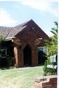 Partial right side view of varied brown brick house with arched porch with lawn in front with the pillar of a similar brown brick fence in the foreground with decorative metal letterbox. 