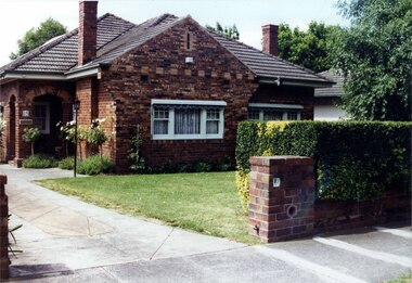 Mixed brown brick house with a double-arched porch and tall feature chimneys with brick patterns plus white windows with a concrete drive, established garden beds and lawn behind a hedge and similar brown brick fence.
