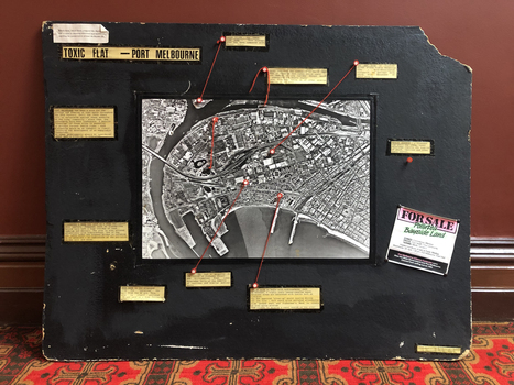 Large board with an aerial photograph of Port Melbourne and a number of captions identifying contaminated sites and connected to the locations in the photograph by red wool.