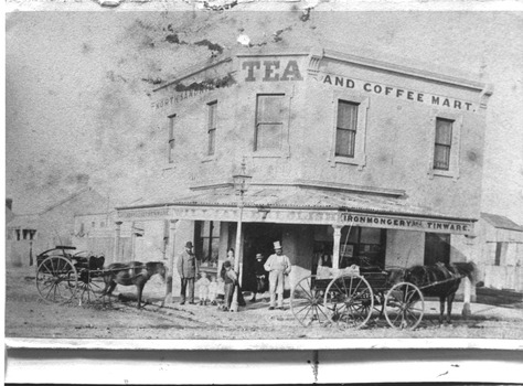 Black & white photo of a two story corner sho[, with horses & carriages on street, several people an also be seen in the photo.