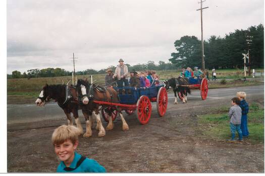 Blue dray with red wheels, full of children, pulled by two dray horses, with one-horse cart behind. Railway crossing in background.