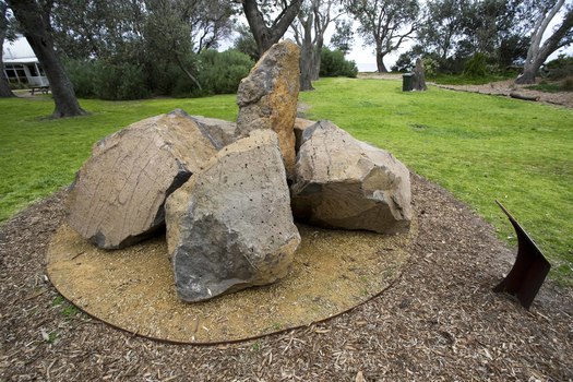 Photograph of an outdoor sculpture comprising of a group of six large bluestones laid on crushed yellow ochre sandstone surrounded by grass and distant vegetation.
