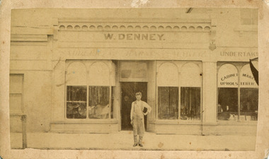 Shop with arched windows either side of door with W. Denny standing on footpath in front