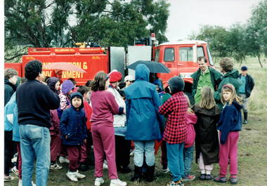 A group of children are gathered around a red truck controlled by the Conservation and Environment personnel while a man explains to the children the importance of the vehicle in their work at the park. 