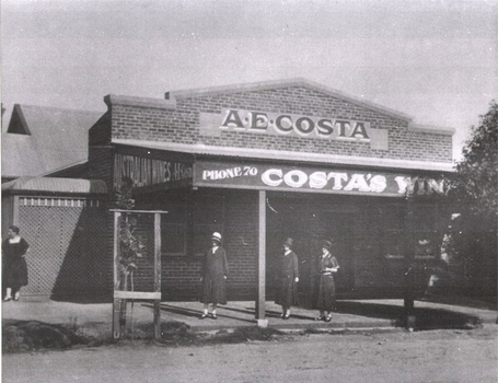 Brick building with advertising signage for A.E. Costa Wine. 4 women are standing outside