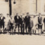 A black and white photograph featuring eleven male figures all dressed in school uniforms standing in a line in-front of a brick building.