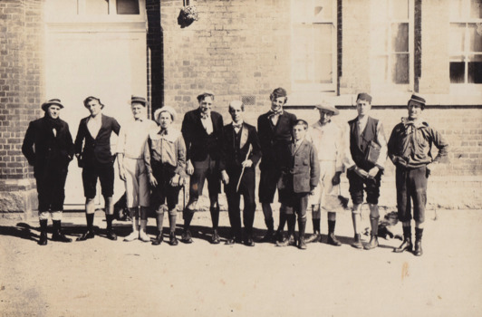 A black and white photograph featuring eleven male figures all dressed in school uniforms standing in a line in-front of a brick building.