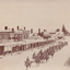 Sepia coloured photograph of a parade of light horsemen riding along a quiet street toward the viewer. The street is lined with businesses, from which a couple of people watch the parade.