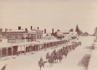 Sepia coloured photograph of a parade of light horsemen riding along a quiet street toward the viewer. The street is lined with businesses, from which a couple of people watch the parade.