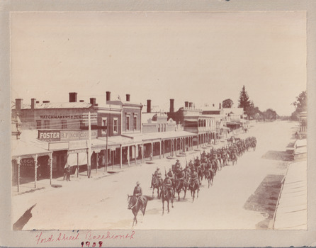 Sepia coloured photograph of a parade of light horsemen riding along a quiet street toward the bottom left of the photo. The street is lined with businesses, from which a couple of people watch the parade. There is writing in red pen along the bottom border.