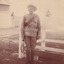 A WWI soldier holding a rifle, standing by a fence. There is a building and fencing in the background.