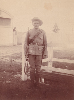 A WWI soldier holding a rifle, standing by a fence. There is a building and fencing in the background.