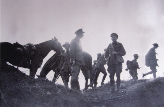 Black and white image of seven World War One soldiers and two horses standing above trenches talking.