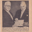 Two men wearing suits standing side-by-side both holding book between them 