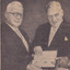 Two men wearing suits standing side-by-side both holding book between them 