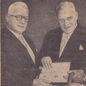 Two men wearing suits standing side-by-side both holding book between them 