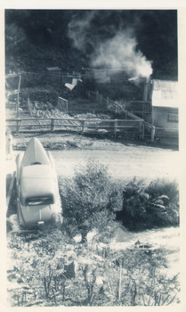 Old car with raised front bonnet beside small bushes and a debris-lined, overgrown shallow crevice. Lane and small cottage with smoking chimney beside a rectangular fenced area. .