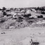 Sloping grassy field with scattered rocks and hillside cutting. A bridge and road leads to a small settlement of scattered buildings. 
