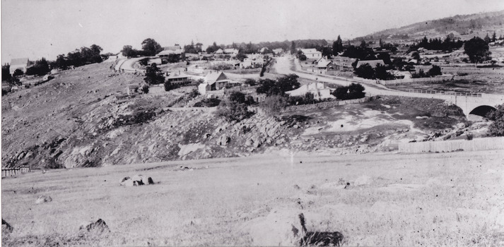 Sloping grassy field with scattered rocks and hillside cutting. A bridge and road leads to a small settlement of scattered buildings. 
