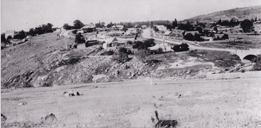 Sloping grassy field with scattered rocks and hillside cutting. A bridge and road leads to a small settlement of scattered buildings. 