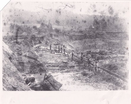 A black and white photograph of a mining landscape and water race with eight individuals standing along the channel. In the background are hills and cottages.