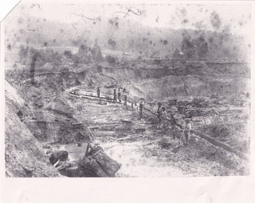 A black and white photograph of a mining landscape and water race with eight individuals standing along the channel. In the background are hills and cottages.