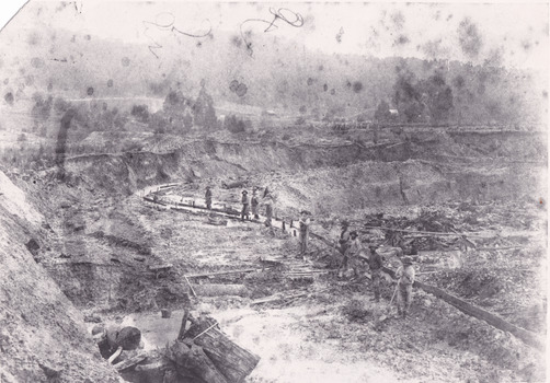 A black and white photograph of a mining landscape and water race with eight individuals standing along the channel. In the background are hills and cottages.