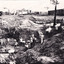 a black and white photograph depicting 22 men in a mining excavation area holding shovels etc. In the background, above the excavation pit, is a row of slab huts.